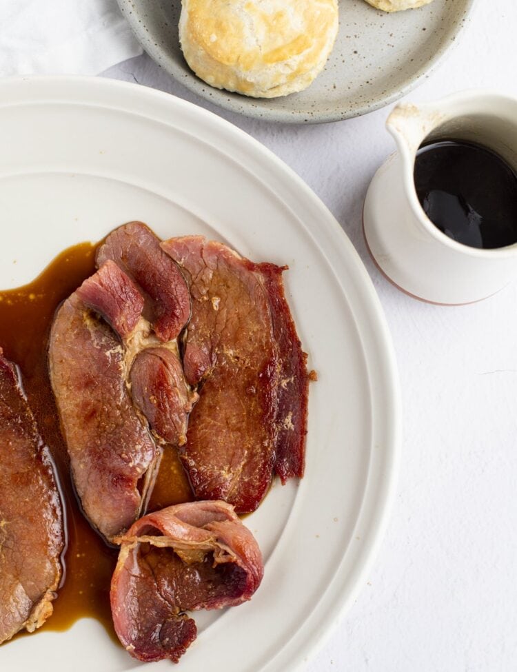 Overhead view of country ham on a white plate on the left side of the photo, with a jar of red eye gravy and a bowl of biscuits above and to the right