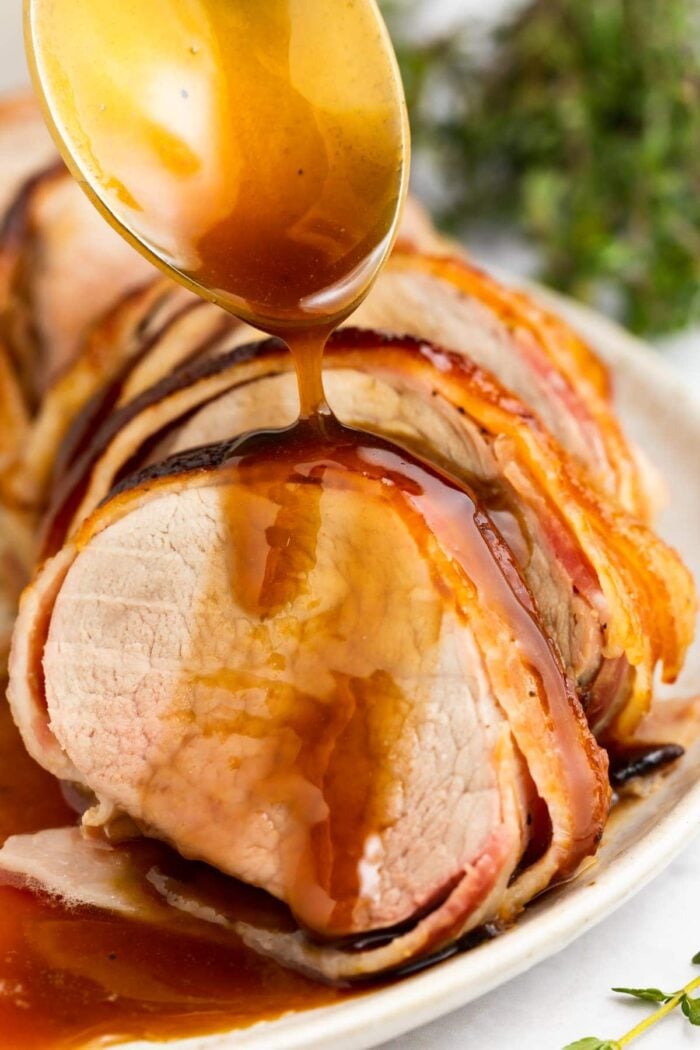 bacon wrapped pork tenderloin on a plate with brown sugar glaze being spooned over the top
