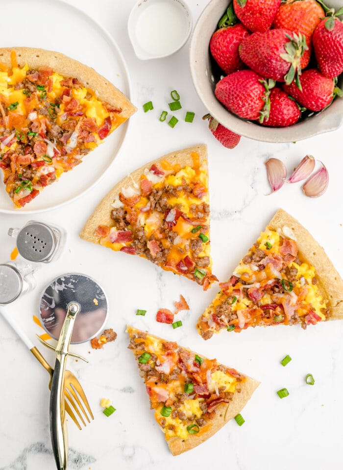 Pieces of breakfast pizza on a countertop with a pizza cutter and a bowl of tomatoes