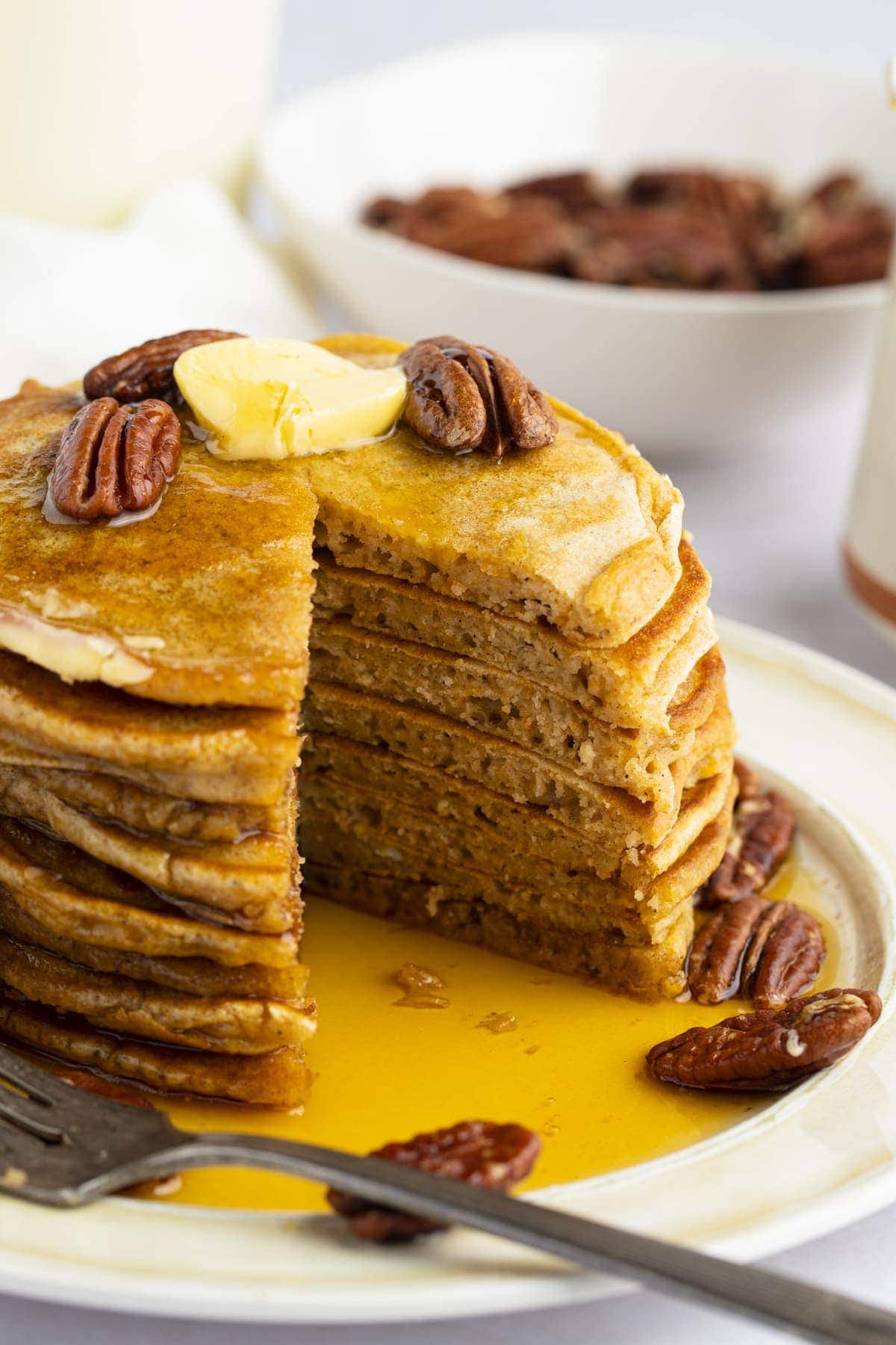 A stack of sweet potato pancakes topped with roasted pecans and butter, with a triangular wedge cut out of the pancakes and resting on a fork.