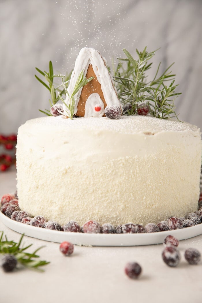 A whole cranberry cake with white chocolate cream cheese frosting on a cake stand, surrounded by loose cranberries