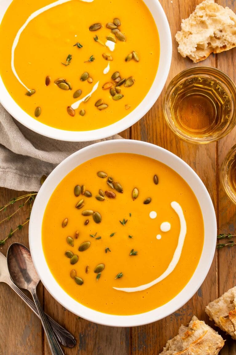 Overhead photo of two white bowls holding creamy, yellow-orange sweet potato soup topped with heavy cream and pumpkin seeds.