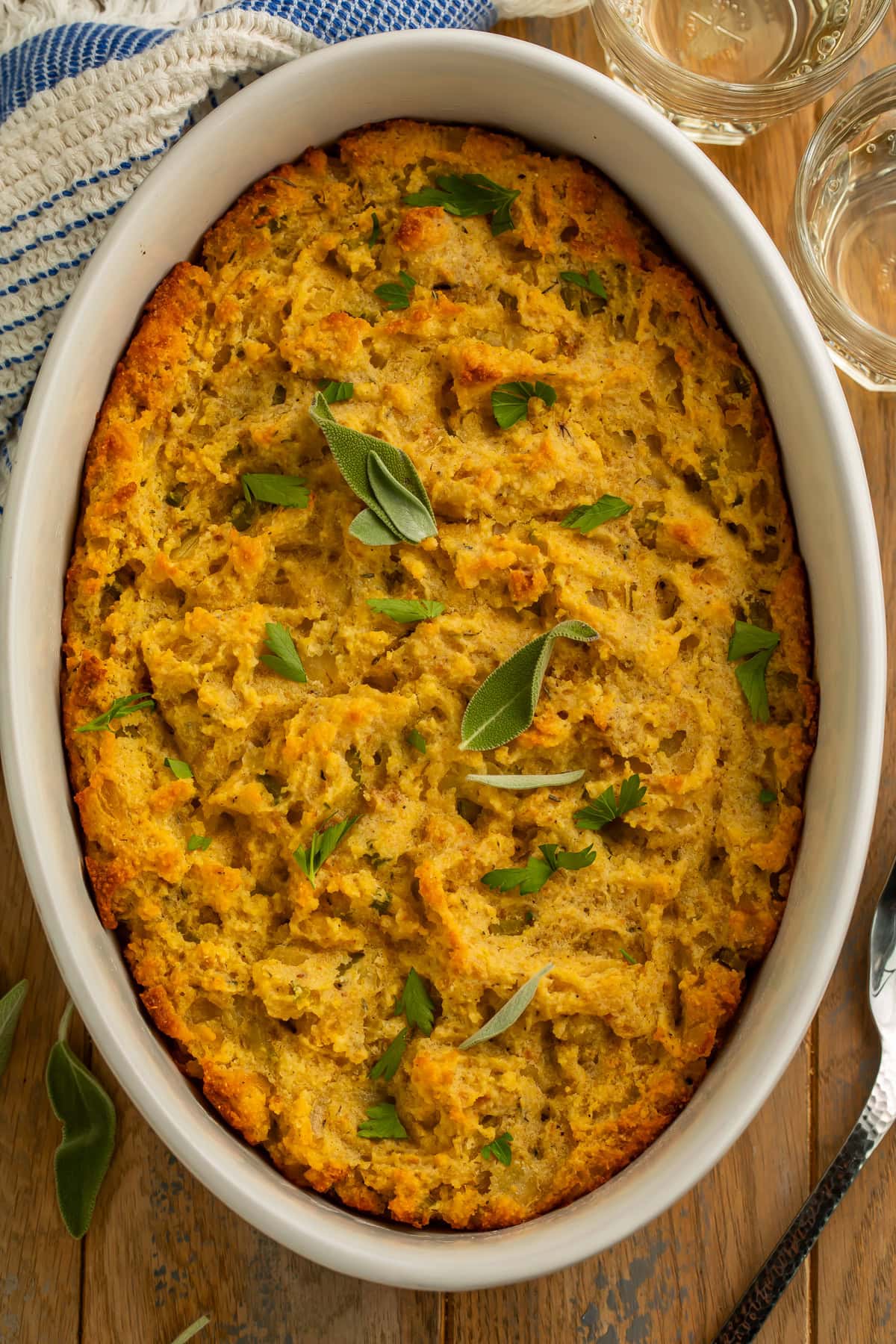 Overhead view of golden-brown cornbread stuffing in an oval baking dish with crispy edges, garnished with sage and parsley.