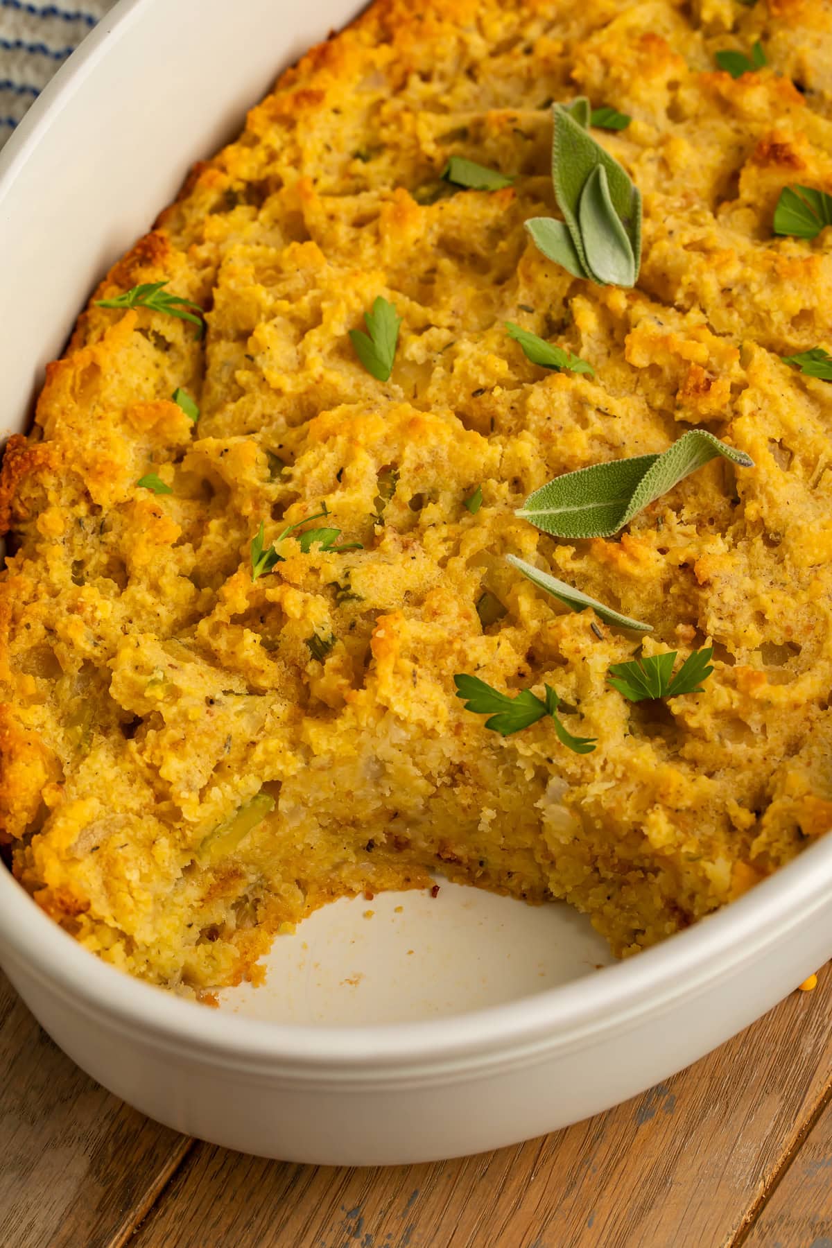 Golden cornbread stuffing in a white oval baking dish with a portion removed, garnished with fresh sage and parsley.