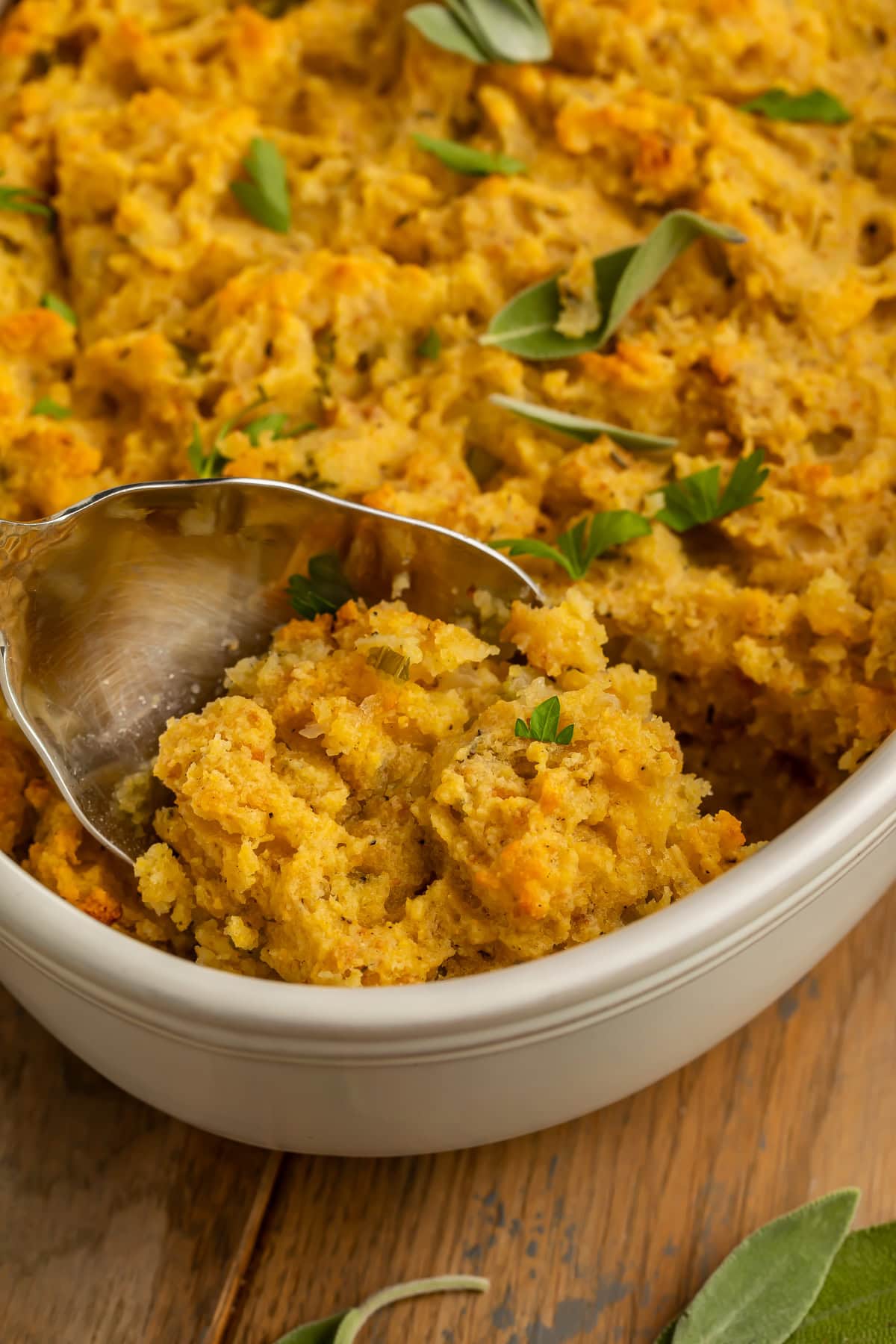 Serving spoon scooping golden cornbread stuffing from a white dish, showing the moist, crumbly texture with fresh herbs.