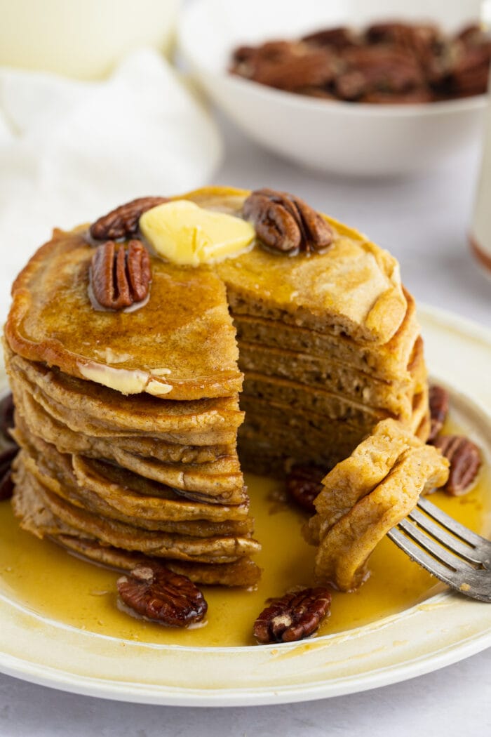 A stack of sweet potato pancakes topped with roasted pecans and butter, with a triangular wedge cut out of the pancakes and resting on a fork