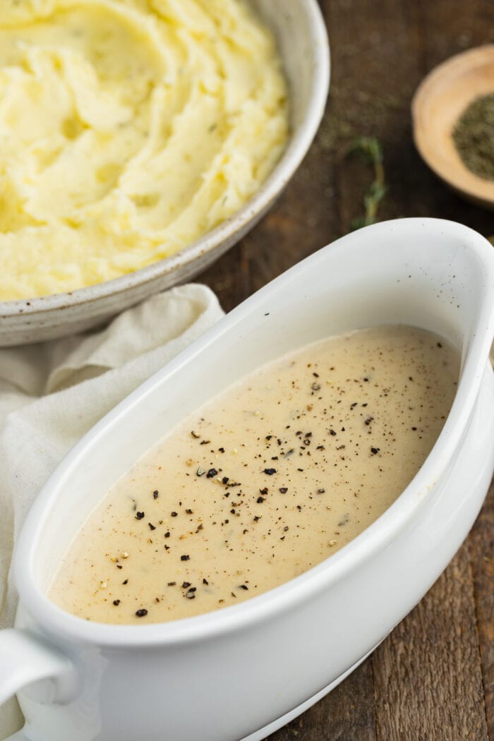 White gravy seasoned with black pepper in a gravy boat next to a bowl of mashed potatoes on a wooden table
