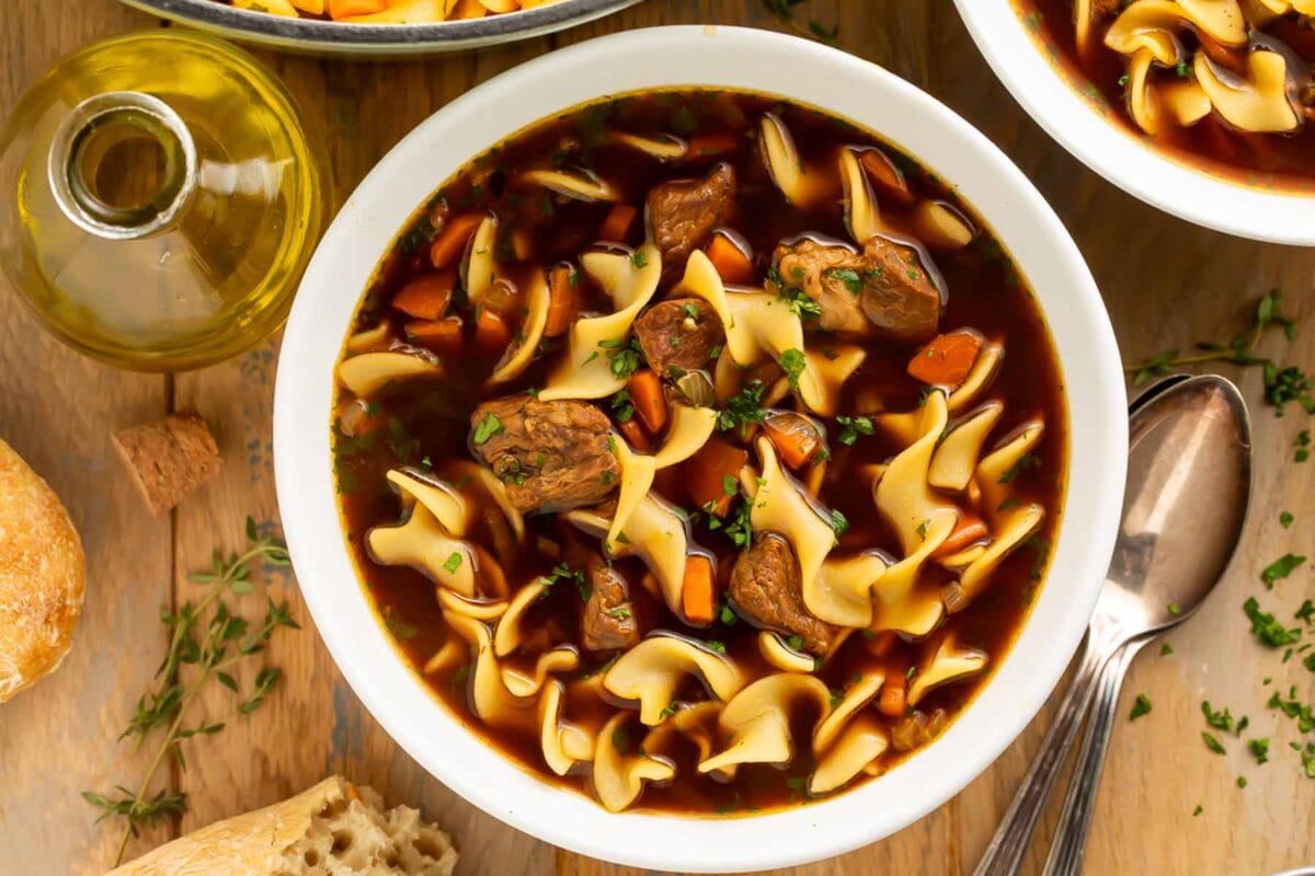 A white soup bowl, holding beef noodle soup with a dark brown broth, sits on a dark wooden tabletop.