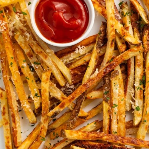 Parmesan truffle fries scattered across a sheet pan lined with parchment paper, next to a ramekin of ketchup.