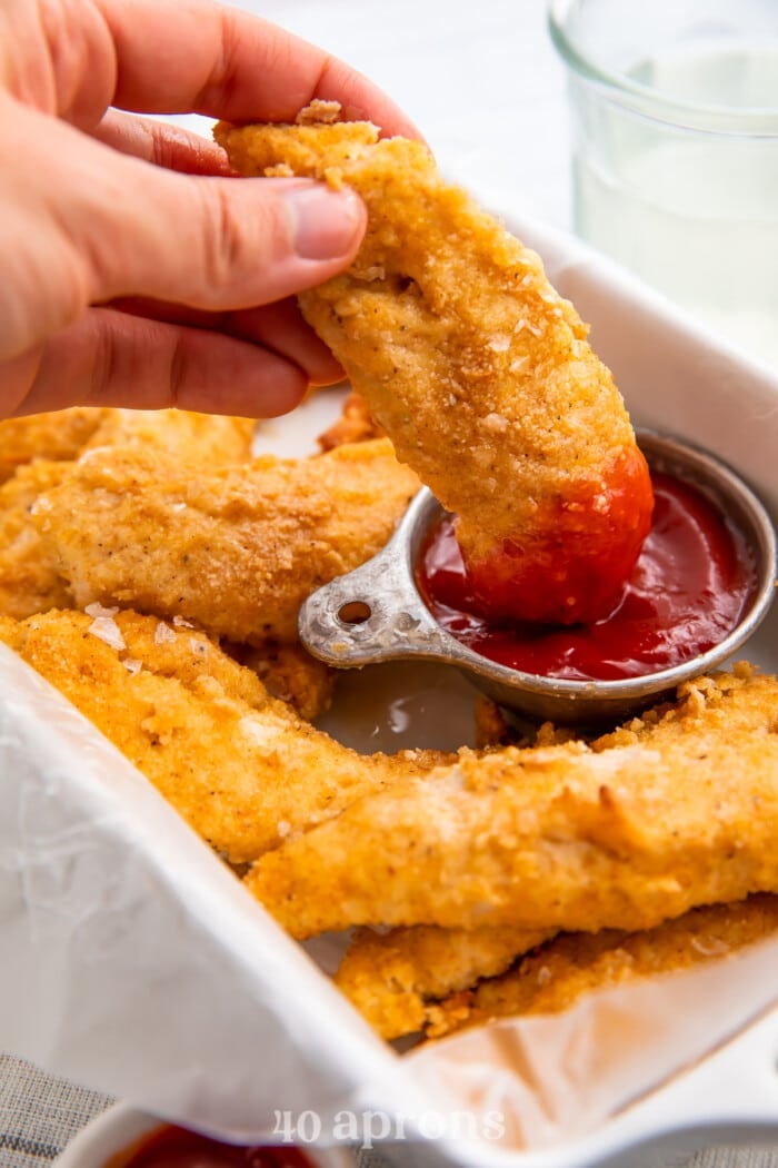 Whole30 chicken tenders surrounding a cup of ketchup while a hand dips a chicken tender into the ketchup
