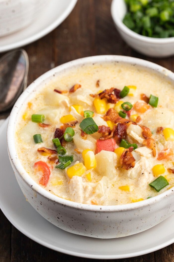 A white bowl of chicken corn chowder sitting on a white plate on top of a dark wooden table.