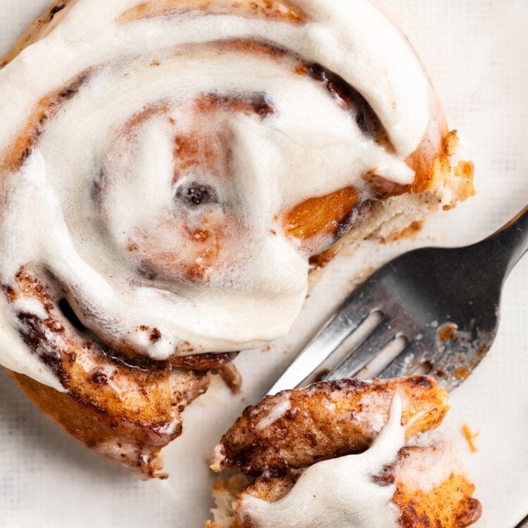 Overhead view of a fork cutting away a piece of a cinnamon roll with cream cheese frosting.