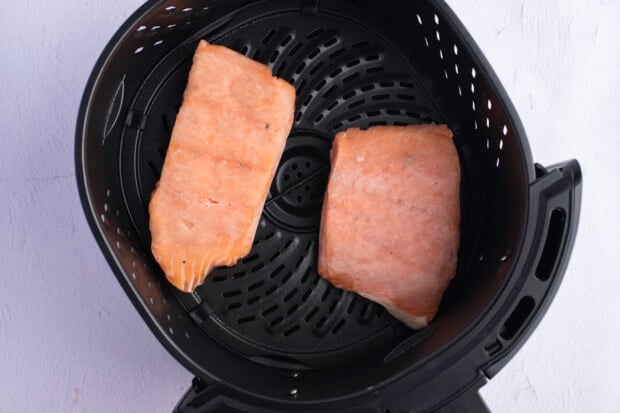 Two 6-ounce frozen salmon fillets in round black air fryer basket on white countertop.
