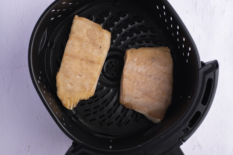 Two 6-ounce frozen salmon fillets in round black air fryer basket on white countertop.