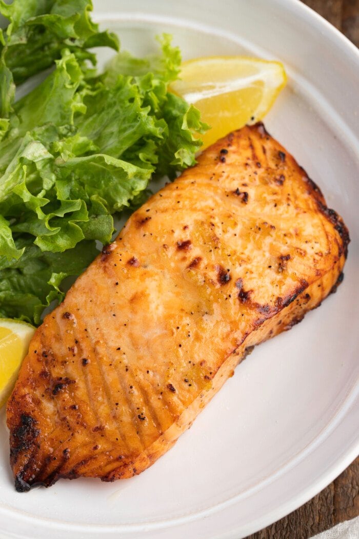 Overhead view of an air fryer frozen salmon fillet on a white plate with green lettuce and lemon wedges.