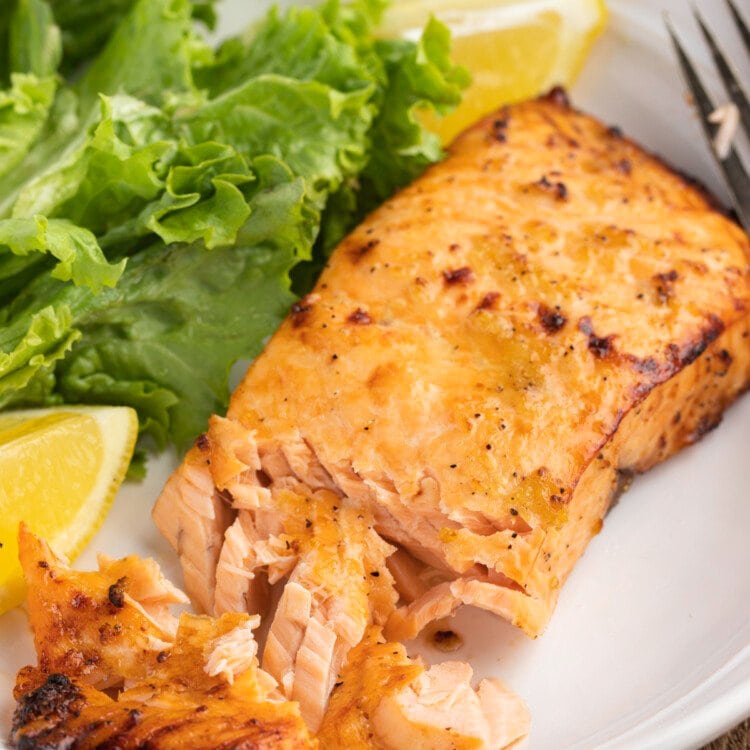3/4-angle view of flaky air fryer salmon fillet on a plate with a small green salad and lemon wedges.