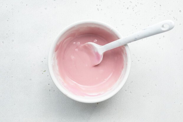 Overhead view of pink vanilla icing for chocolate gluten-free donuts in a large glass mixing bowl with a spoon.