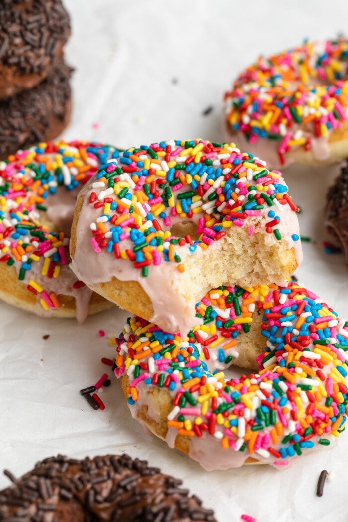 3/4 angle photo of a stack of vanilla donuts with rainbow sprinkles, with one bite taken out of one donut.