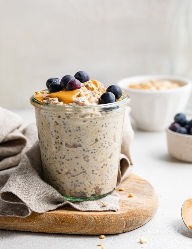 A glass of peanut butter overnight oats topped with peanut butter and blueberries on a wooden serving board.