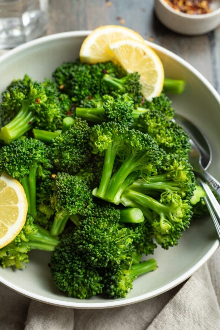 Overhead view of a bowl of steamed broccoli florets with leon wedges on a wooden tabletop.