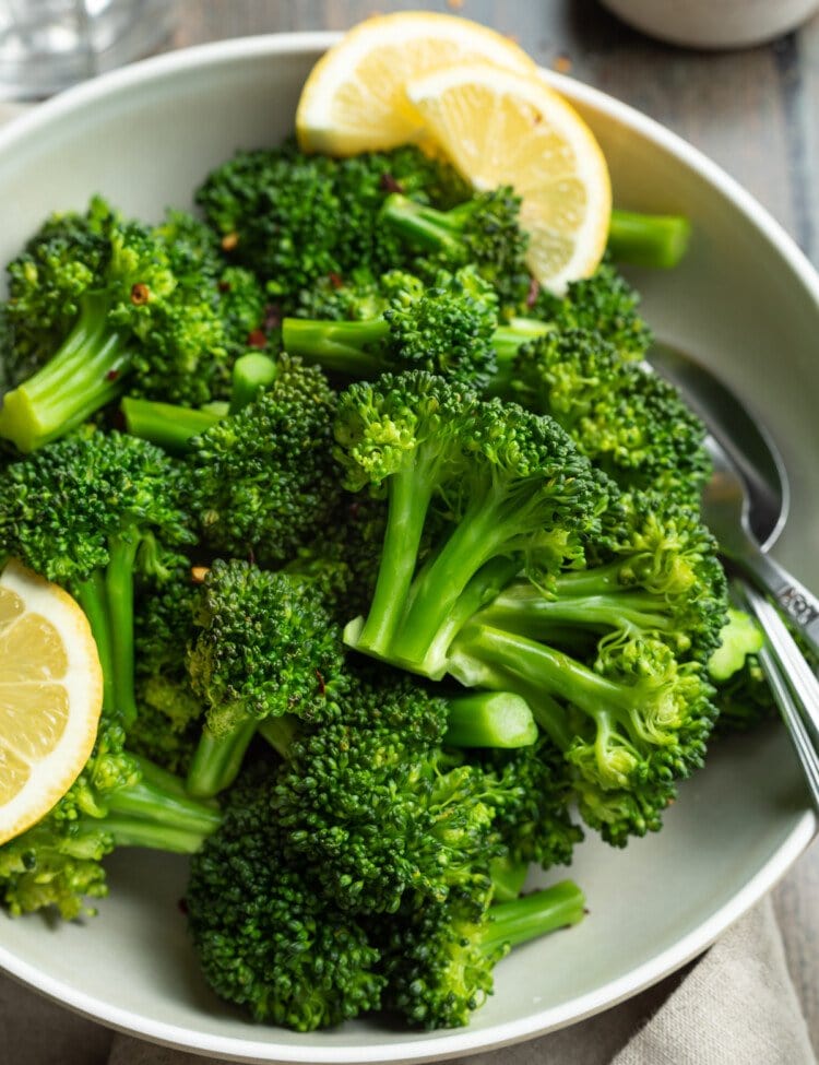 Overhead view of a bowl of steamed broccoli florets with leon wedges on a wooden tabletop.