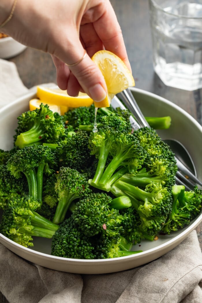 A hand squeezing juice from a lemon wedge over a bowl of green steamed broccoli florets on a wooden table.