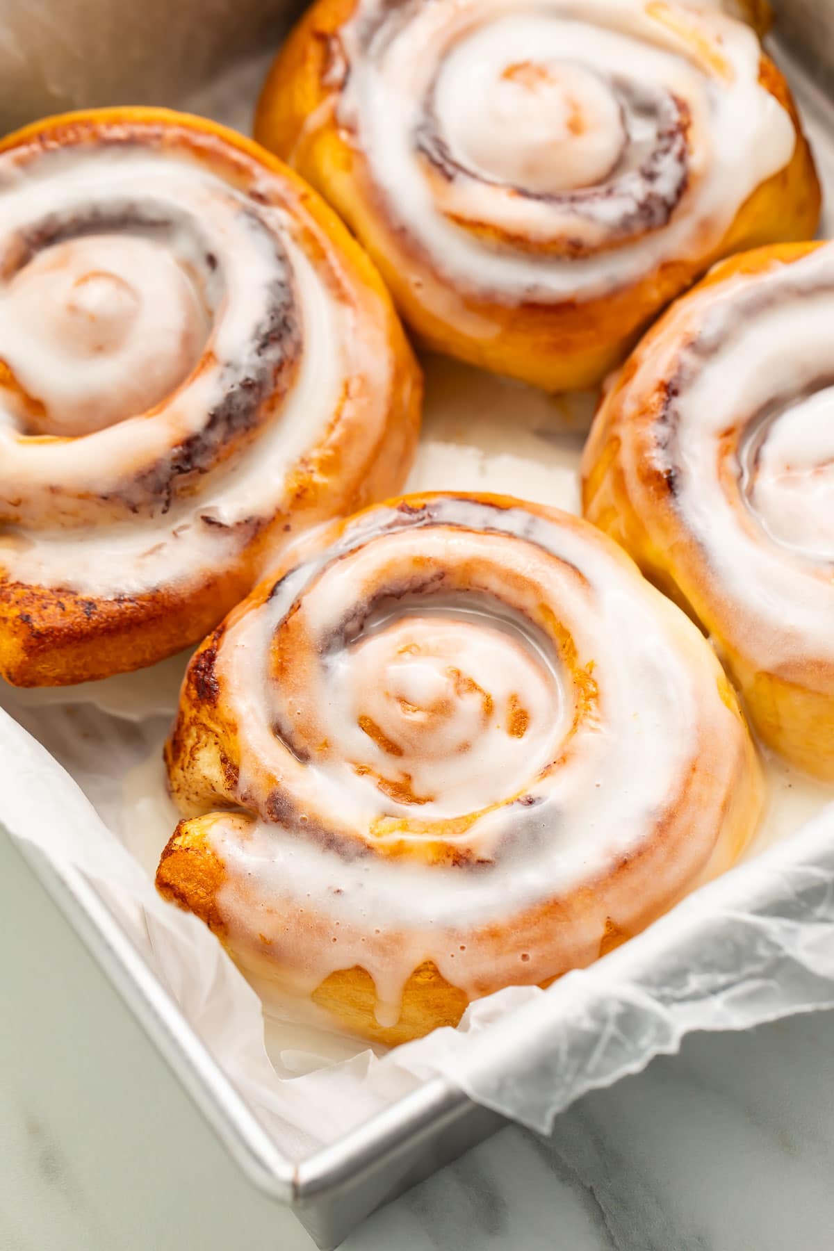 4 air fryer cinnamon rolls topped with melty cinnamon roll icing resting in a pan lined with parchment paper.
