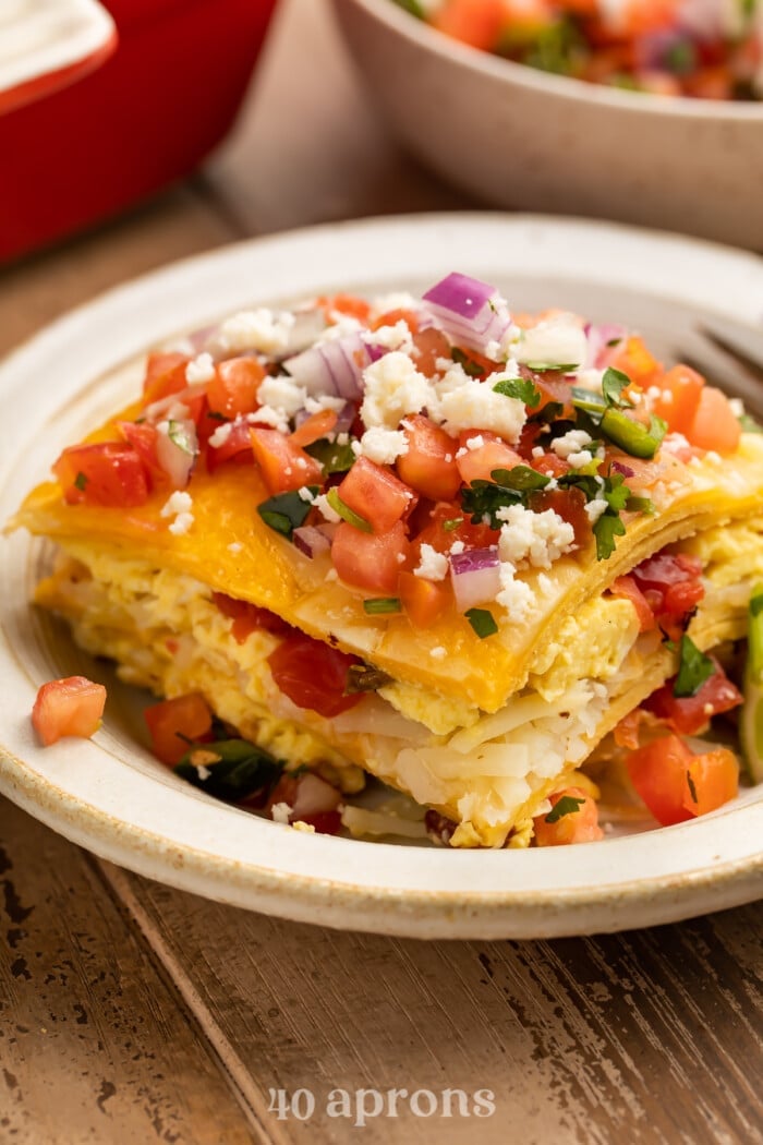 A square slice of breakfast taco casserole on a white plate, showing the layers of tortilla, eggs, and pico.