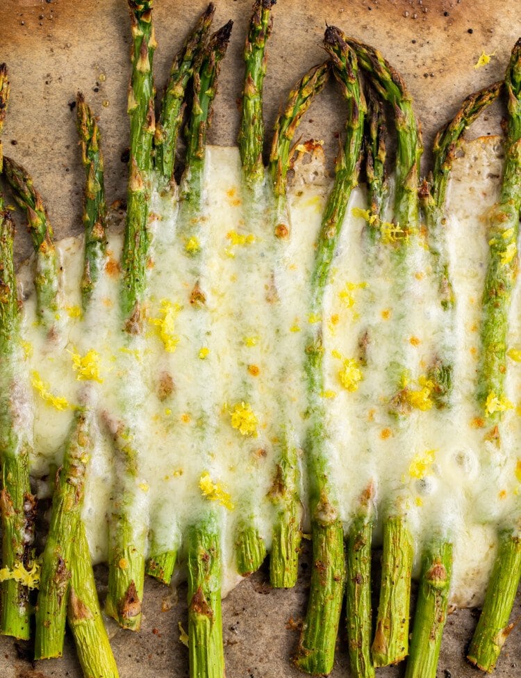 Overhead view of roasted asparagus spears covered in cheese on a baking sheet lined with parchment paper.