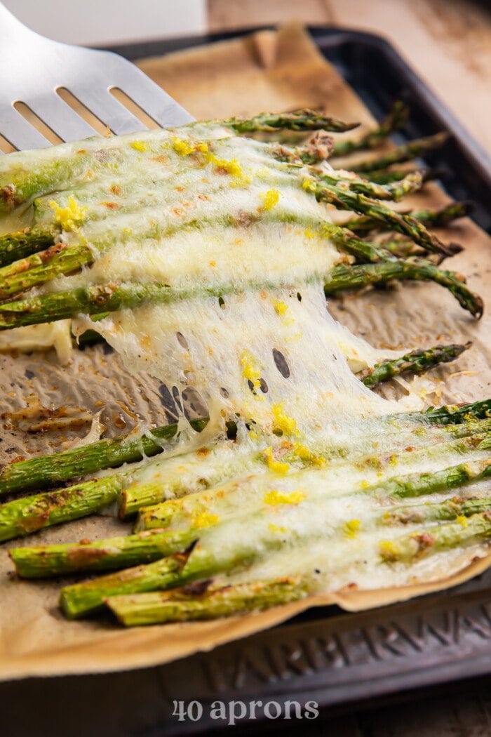 A portion of roasted asparagus is lifted off a baking sheet by a metal spatula, resulting in a cheese pull.
