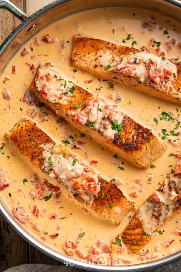Overhead view of 4 salmon fillets in a large silver saucepan with red pepper parmesan sauce.