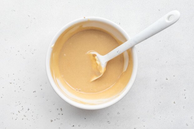 Overhead view of a small bowl of honey mustard sauce with a white silicone spatula on a white background.