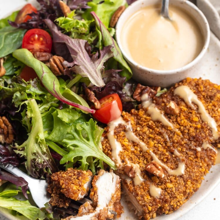 Close-up, angled view of pecan crusted chicken with honey mustard sauce and small side salad on a white plate.
