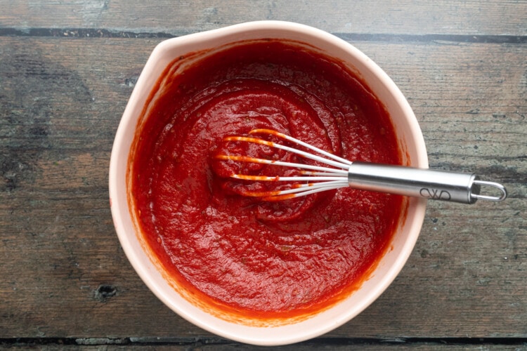 Overhead view of bright red pizza sauce in a white mixing bowl with a whisk.