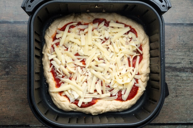 Overhead view of an unbaked air fryer pizza topped with pizza sauce and shredded cheese in an air fryer.