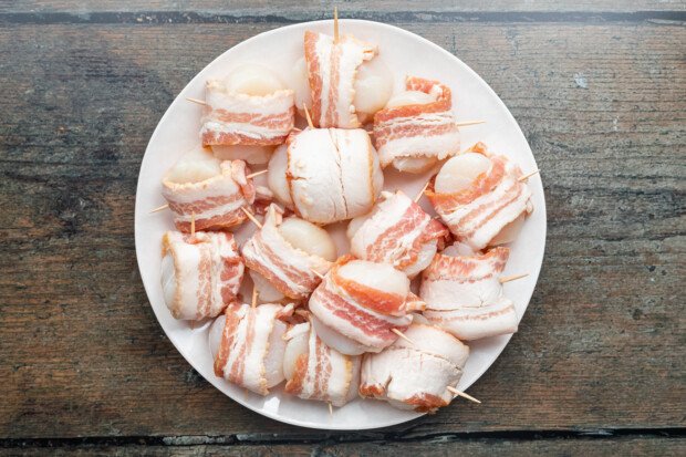 Bacon-wrapped scallops, uncooked, on a white plate.