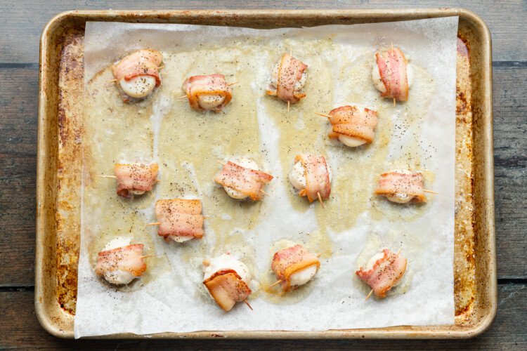 Bacon-wrapped scallops drizzled with olive oil, salt, and pepper, on a baking sheet lined with parchment paper.