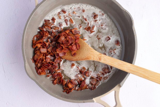 Overhead view of chopped bacon pieces in large grey skillet with wooden spoon and rendered bacon fat.