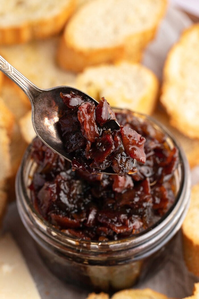 Overhead, 3/4-angle view of a spoonful of bacon jam being lifted out of a jar, with crostini in the background.