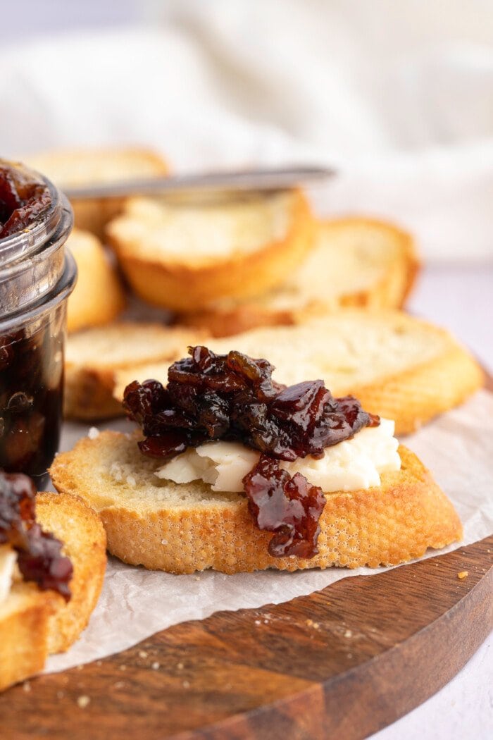 Side view of bacon jam atop crostini and cheese on a wooden cutting board.