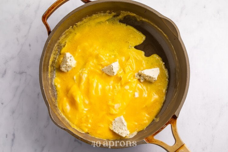 Overhead view of a large skillet with barely-set scrambled eggs and chunks of Boursin cheese.