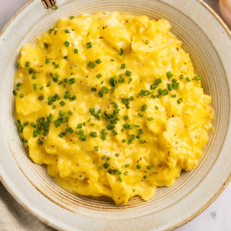 Overhead view of a bowl of boursin scrambled eggs garnished with chives on a neutral countertop.