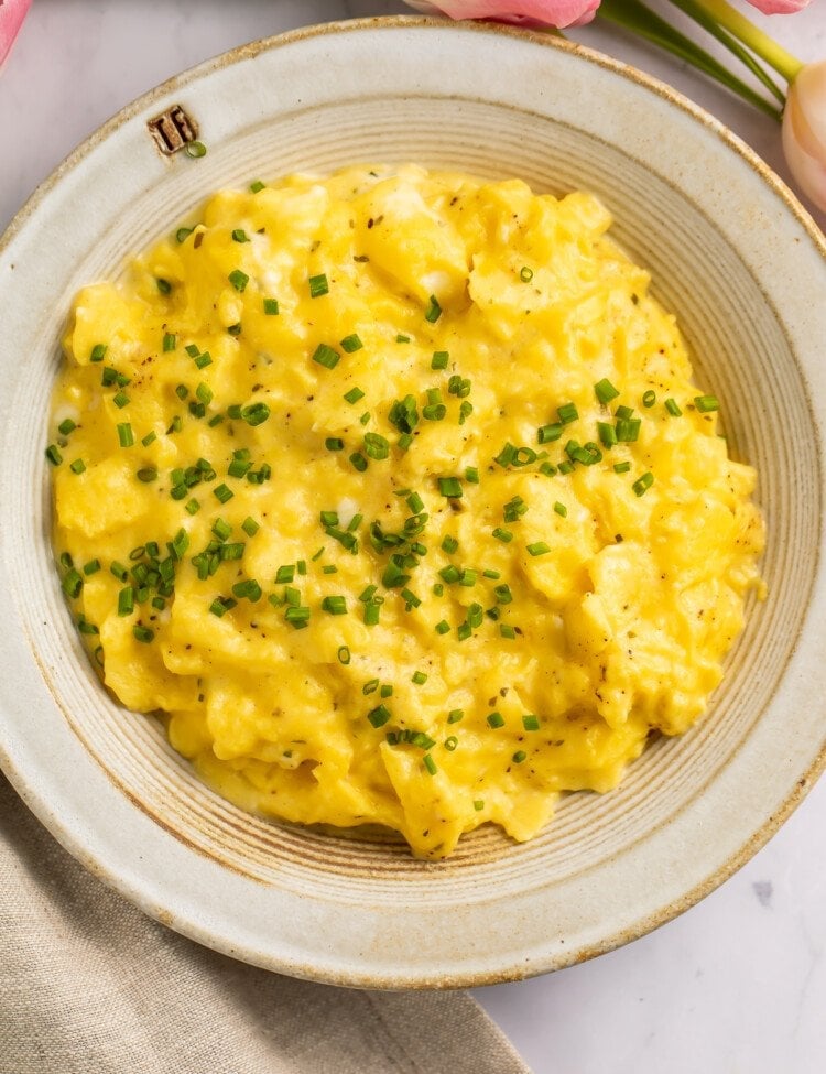 Overhead view of a bowl of boursin scrambled eggs garnished with chives on a neutral countertop.