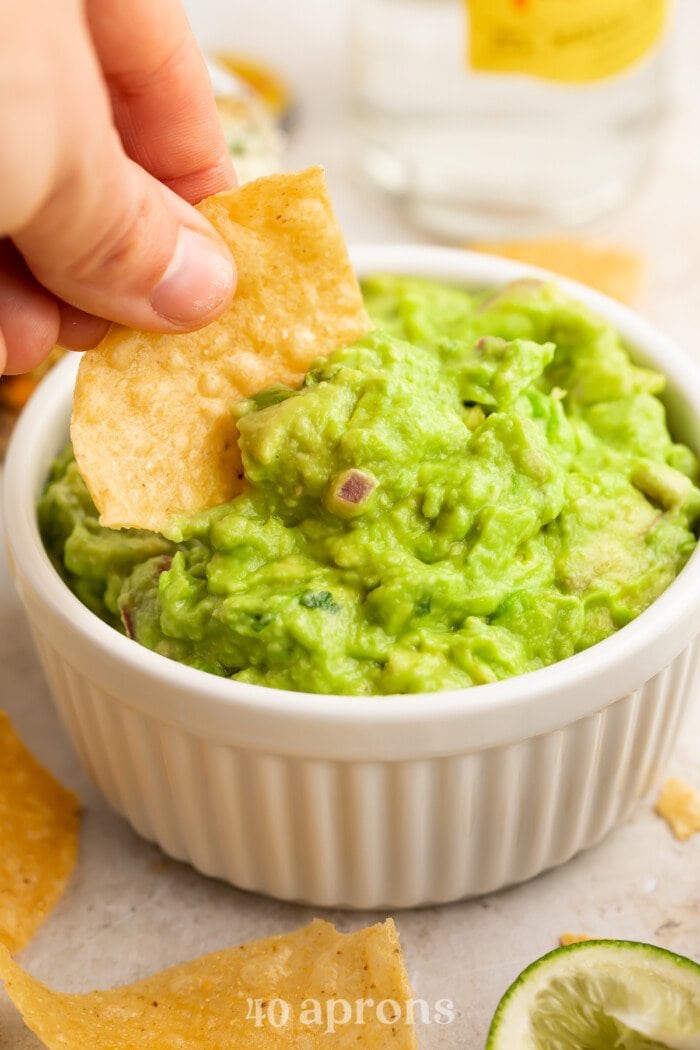 A woman's hand dipping the corner of a tortilla chip in a ramekin of Chipotle guacamole with onion and jalapeno.
