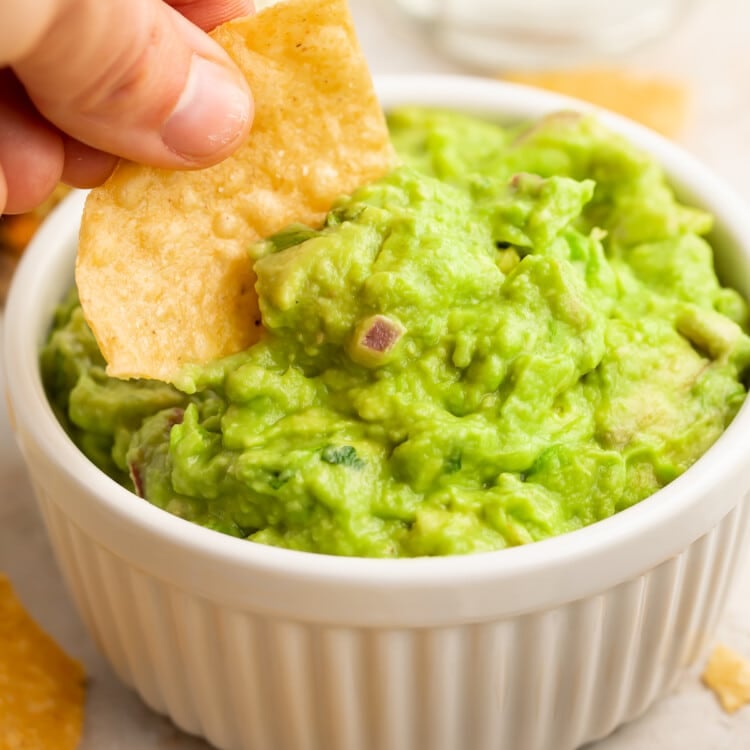 A woman's hand dipping the corner of a tortilla chip in a ramekin of Chipotle guacamole with onion and jalapeno.