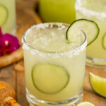 3/4-angle overhead view of a rimmed glass with cucumber margarita and lime wedges.