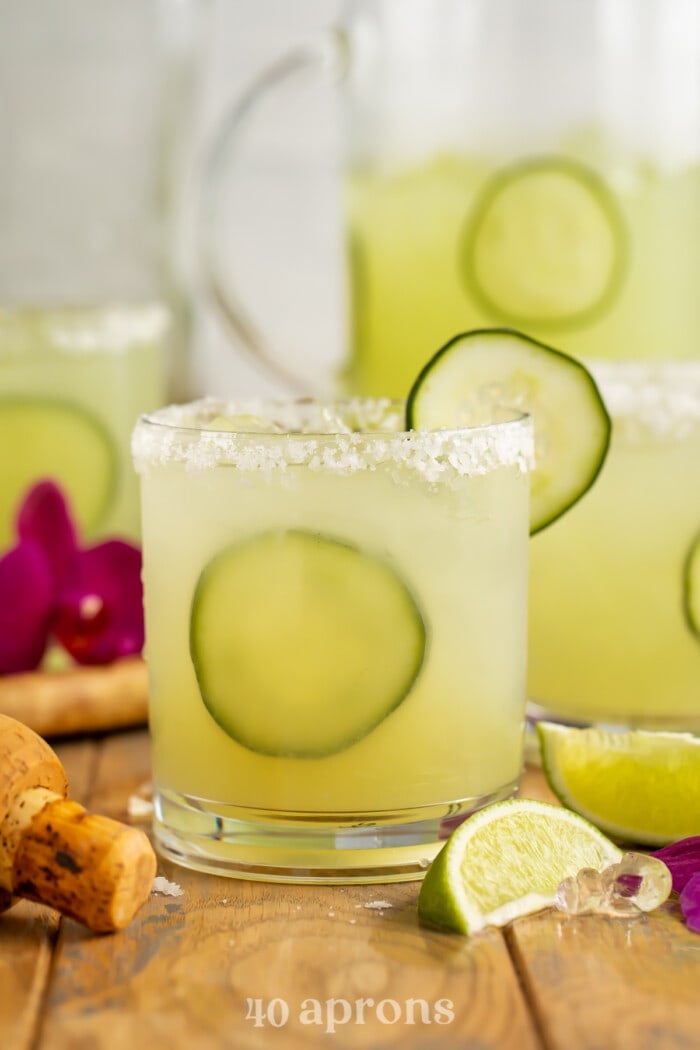 Side view of a glass of cucumber margarita with cucumber garnish in front of a pitcher of margaritas.