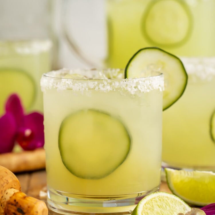 Side view of a glass of cucumber margarita with cucumber garnish in front of a pitcher of margaritas.