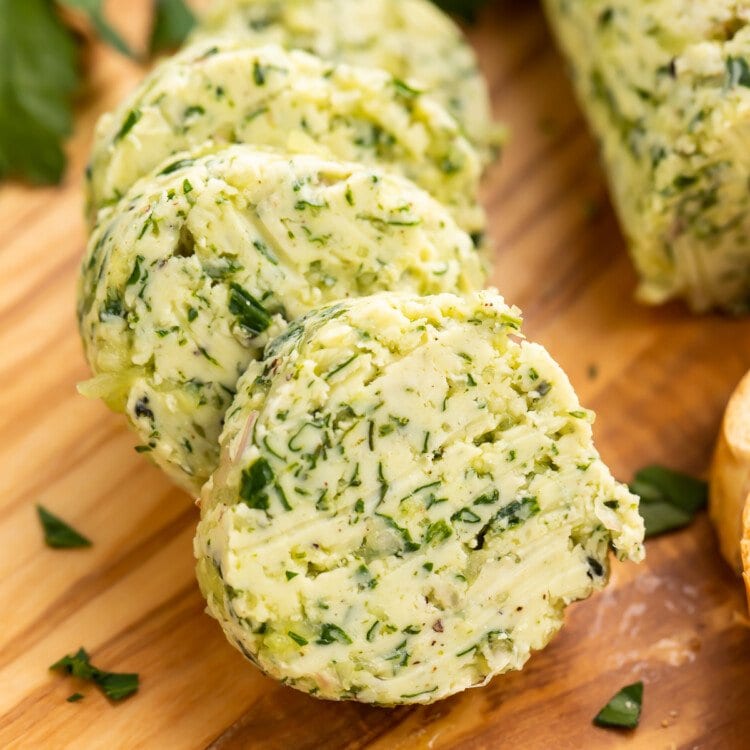 Close-up view of a log of garlic herb butter sliced into discs on a wooden serving board.