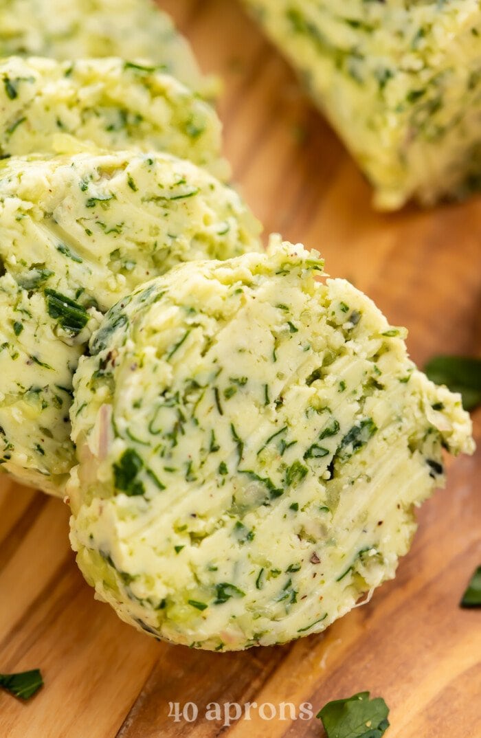 Close-up view of a log of garlic herb butter sliced into discs on a wooden serving board.